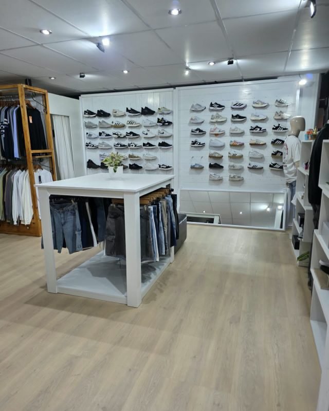 Interior del local SCH: pared blanca llena de zapatillas en repisas, mesa blanca con jeans colgados y perchero de madera con ropa