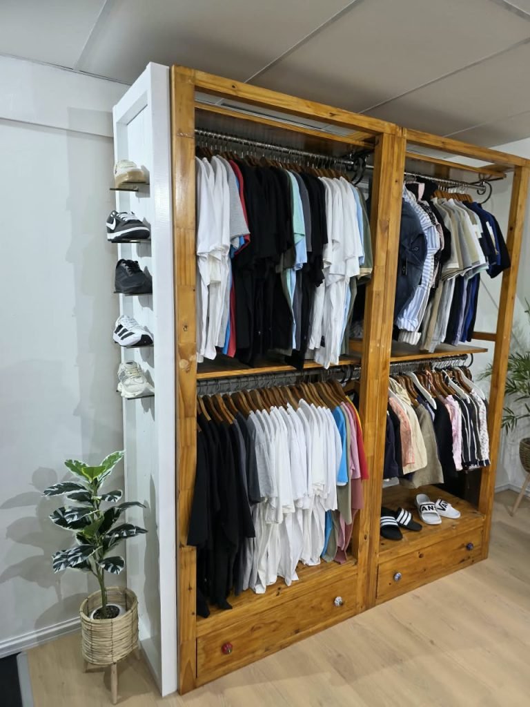 Perchero de madera del local SCH con remeras, camisas y ojotas, y una repisa con zapatillas al costado
